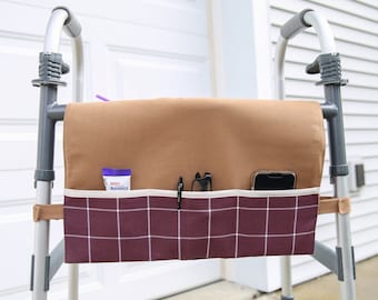 May include: A brown and white fabric organizer with multiple pockets attached to a silver walker. The organizer has a brown top flap and a plaid bottom section with a white trim. The pockets contain a tube of hand cream, a pair of glasses, and a cell phone.