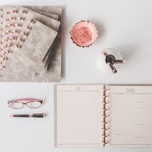 May include: A set of three beige faux leather journals with gold rings and a rose gold pen. The journals are stacked on top of each other and have a subtle crocodile pattern. The pen is lying on top of the journals. There is a small pink dish with a floral design and a white ceramic holder with three rose gold pens.