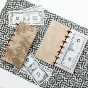 May include: A beige leather-bound planner with a gold ring binding. The planner is open and has a dollar bill tucked into the left side. The planner is on a black and white patterned surface.