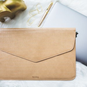 May include: A tan laptop sleeve with a crocodile texture, a gold pen, and paper clips are on a white surface. A gold sculpture and a silver laptop are also visible. The brand name "NOTIQ" is printed on the sleeve.