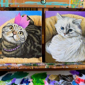 May include: Two small square paintings of cats. One shows a tabby cat wearing a pink crown and a purple necklace. The other painting depicts a fluffy white cat with blue eyes. Both cats are painted on a canvas with a purple and yellow background.