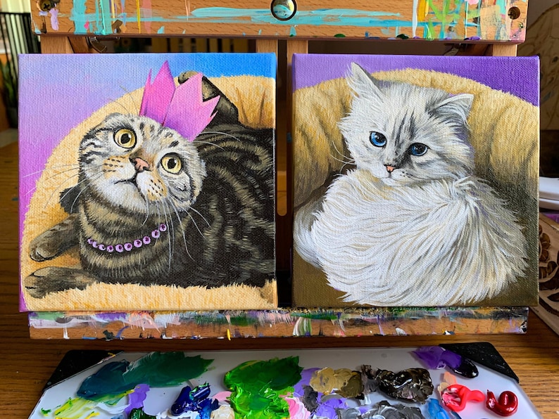 May include: Two small square paintings of cats. One shows a tabby cat wearing a pink crown and a purple necklace. The other painting depicts a fluffy white cat with blue eyes. Both cats are painted on a canvas with a purple and yellow background.