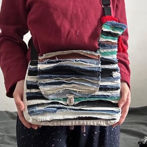 Puede incluir: Un bolso bandolera de mezclilla de retazos coloridos con un diseño único y superpuesto. El bolso tiene una solapa de cierre con una hebilla plateada y una correa larga. El bolso está decorado con un pequeño amuleto de animal de crochet colorido.