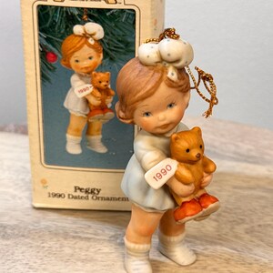 May include: A porcelain Christmas ornament of a girl holding a teddy bear, dated 1990. The girl wears a light blue dress, white socks, and orange shoes. The ornament is from the "Memories of Yesterday" collection.