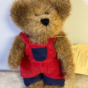 May include: A brown teddy bear wearing red corduroy overalls with a navy blue pocket and navy blue cuffs. The bear has a black nose and a tag attached to its side. The plush toy is a classic children's item.