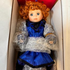 May include: A porcelain doll with red curly hair and blue eyes, wearing a blue dress with black trim. The doll is packaged in a cardboard box with a red interior, protected by bubble wrap.