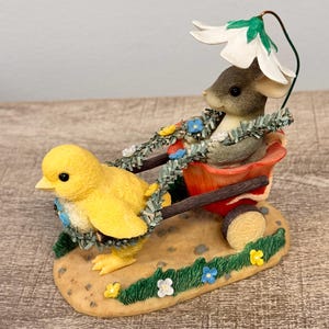 May include: A whimsical figurine featuring a yellow chick pulling a red wagon with a gray mouse inside. The chick and mouse are adorned with floral garlands. A large white flower provides shade. The base is decorated with grass and flowers.