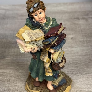 May include: A figurine of a woman in a green dress, holding stacks of books and papers. She wears glasses on her head and carries a small bag. The base reads "Teaching is Anything but Elementary!"