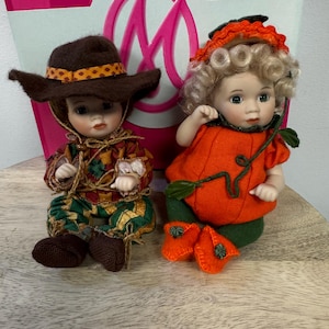 May include: Two porcelain dolls. One doll is dressed as a scarecrow in a brown hat and patchwork clothing. The other doll is dressed as a pumpkin with an orange outfit, green leggings, and curly blonde hair. Both dolls are on a wooden surface.