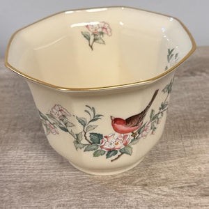 May include: Cream-colored ceramic bowl with a gold rim and a floral and bird design. The bowl features a red bird perched on a flowering branch with pink and white blossoms. The bowl has an octagonal shape.