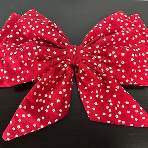 May include: A large fabric hair bow in a vibrant red color featuring a repeating pattern of tiny white flowers and small black dots. The bow has a structured, multi-layered design with two long, pointed tails extending downward from the center knot.