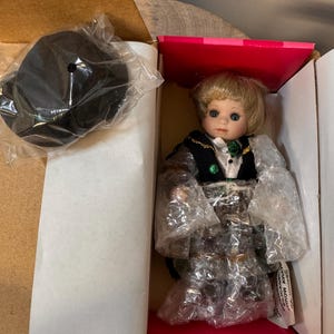 May include: A porcelain doll with blonde hair and blue eyes, wearing a black and green vest over a white shirt, packaged in a box. A black stand is included. The doll is wrapped in plastic.