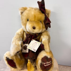 May include: A tan teddy bear with a burgundy bow, a burgundy pouch, and a gold tassel. The bear has burgundy accents on its paws and the words "Cherished Teddies" are printed on the paws. A tag reads "Love is the thread that pulls our hearts and souls together."