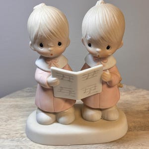 May include: A porcelain figurine of two children singing from a music book. They are wearing pink dresses and have blonde hair. The figurine is on a white base.