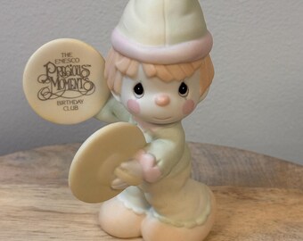 May include: A Precious Moments figurine of a child wearing a hat and holding two cymbals. The figurine is in pastel colors with the text "The Enesco Precious Moments Birthday Club" on one cymbal.