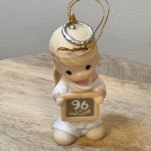 May include: A ceramic figurine of a kneeling angel holding a small chalkboard. The angel wears a white robe with pink accents and has blonde hair. The chalkboard reads "96 Peace on Earth" in brown lettering, and has a gold string for hanging.