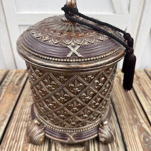 May include: A decorative lidded jar featuring a dark brown and metallic silver finish with a raised fleur-de-lis pattern in a diamond grid. The lid has a finial top and a black rope tassel attached. The jar stands on three ornate, curved feet.