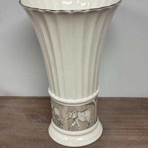 May include: A cream-colored ceramic vase with a fluted, scalloped rim and a silver-toned edge. The base features a decorative band with a detailed black and white illustration of elephants walking through a grassy landscape against a tan background.