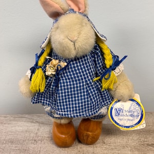 May include: A plush bunny doll wearing a blue and white checkered dress, a matching bonnet, and wooden shoes. The bunny has yellow yarn braids and a tag that reads "Muffy VanderBear Dutch Treat".