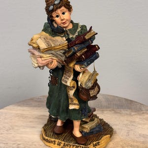 May include: A figurine of a woman in a green dress, holding a stack of papers and books, and a phone to her ear. The base of the figurine reads "Teaching is Anything but Elementary!"