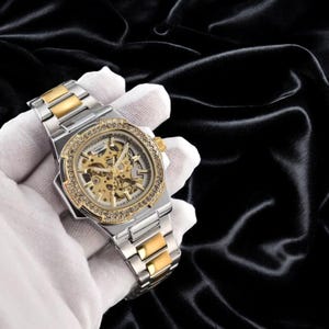 May include: A two-tone silver and gold stainless steel wristwatch featuring a skeleton dial that reveals the internal mechanical movement. The round bezel is encrusted with clear crystals, and the metal link bracelet has a folding clasp.
