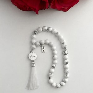 K&ouml;nnte beinhalten: Wei&szlig;e Gebetsperlen mit einem wei&szlig;en Quasten und einem silbernen Anh&auml;nger mit dem arabischen Wort "أحبك" (Ich liebe dich) und dem Buchstaben "N".