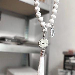 K&ouml;nnte beinhalten: Armband aus wei&szlig;en Marmorperlen mit einem silbernen Anh&auml;nger, auf dem "Demir" steht, und einem wei&szlig;en Quasten.
