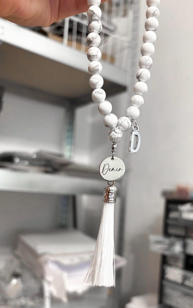 K&ouml;nnte beinhalten: Armband aus wei&szlig;en Marmorperlen mit einem silbernen Anh&auml;nger, auf dem "Demir" steht, und einem wei&szlig;en Quasten.
