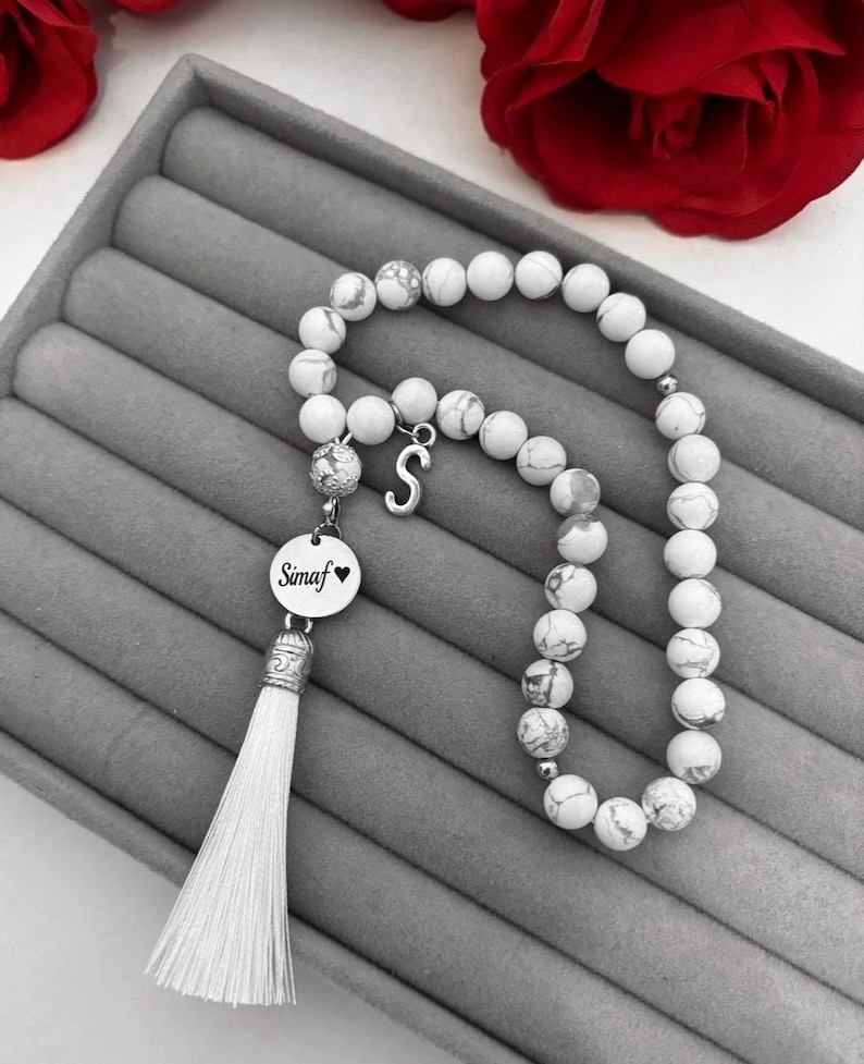 K&ouml;nnte beinhalten: Armband aus wei&szlig;en Marmorperlen mit einem silbernen Anh&auml;nger, auf dem "Simaf" mit einem Herzen steht. Das Armband hat einen wei&szlig;en Quasten.