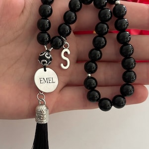 K&ouml;nnte beinhalten: Schwarzes Perlenarmband mit einem silbernen Anh&auml;nger, auf dem "EMEL" steht, und einem silbernen Anh&auml;nger mit dem Buchstaben "S". Das Armband hat einen schwarzen Quasten.