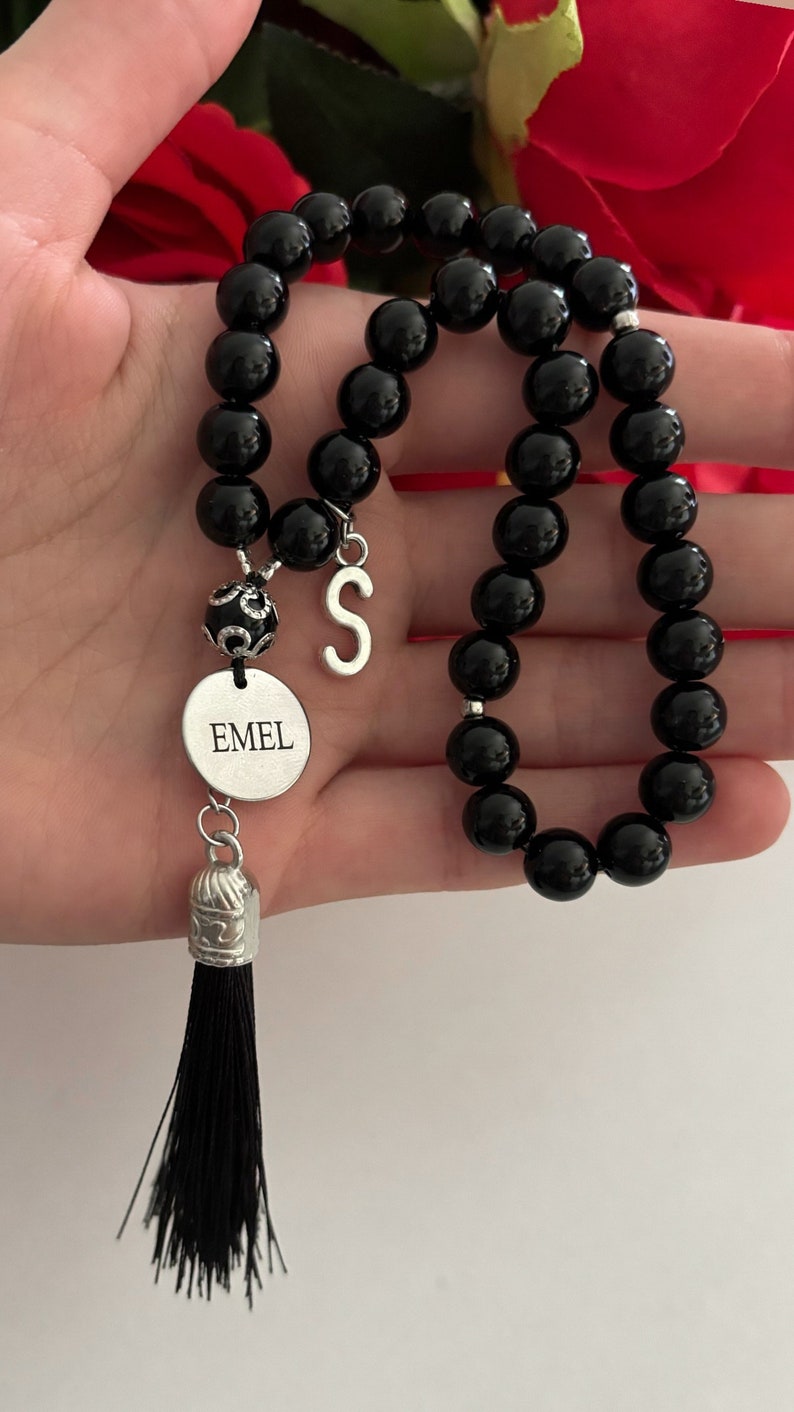 K&ouml;nnte beinhalten: Schwarzes Perlenarmband mit einem silbernen Anh&auml;nger, auf dem "EMEL" steht, und einem silbernen Anh&auml;nger mit dem Buchstaben "S". Das Armband hat einen schwarzen Quasten.