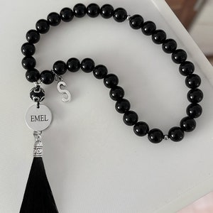 K&ouml;nnte beinhalten: Schwarze Gebetsperlen mit einem silbernen Quasten und einem Anh&auml;nger mit dem Namen "EMEL" und dem Buchstaben "S".