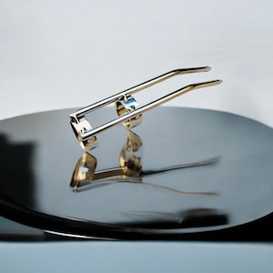 Può includere: Un tagliaunghie in metallo argentato dal design curvo, appoggiato su una superficie circolare riflettente. Il tagliaunghie presenta due barre parallele e un tagliente. La superficie riflette la forma del tagliaunghie, creando un'immagine speculare.