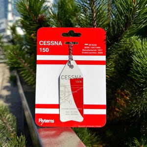 May include: A Cessna 150 keychain on a red and white backing card. The keychain is silver with the Cessna 150K logo and a red and white design. The card includes the text "CESSNA 150" and "Flytems".