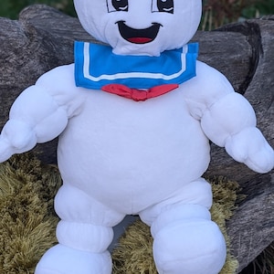 Op de afbeelding: Een wit pluche speelgoed van de Stay Puft Marshmallow Man uit Ghostbusters. Het speelgoed draagt een blauwe matrozenkraag met een rode strik en een witte matrozenpet met de tekst "Stay Puft" erop. Het speelgoed zit op een groene, pluche ondergrond.