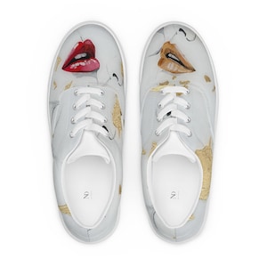 May include: A pair of light gray lace-up sneakers featuring a graphic print of red lips on the left shoe and gold lips on the right shoe. The design includes scattered gold leaf accents across the surface and the letters ND printed on the insoles.