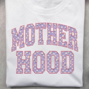 Op de afbeelding: Wit T-shirt met het woord "MOTHERHOOD" in een paars bloemenpatroon. De tekst is in een lettertype in collegestijl met een roze omlijning. Het shirt is op een grijze ondergrond gevouwen.