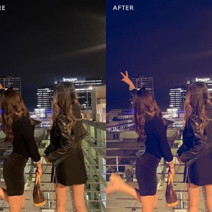 May include: Two people pose for a photo in front of a city skyline at night. They are both wearing black outfits and have their arms raised in the air. The city lights are visible in the background.