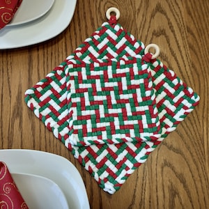 Puede incluir: Dos agarraderas de olla tejidas en rojo, blanco y verde con asas de madera. Las agarraderas están apiladas una encima de la otra.