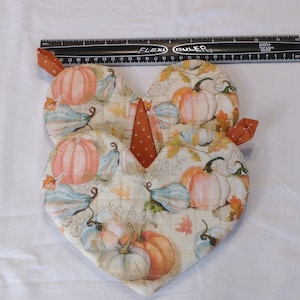 May include: Two heart-shaped quilted pot holders with a fall theme. The pot holders feature a cream background with a pattern of pumpkins, gourds, and leaves in shades of orange, brown, and green.