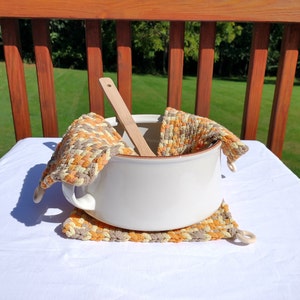 May include: A white ceramic pot with a wooden spoon inside, sitting on two woven pot holders with a brown, orange, and cream color pattern. The pot holders have loops on the sides.