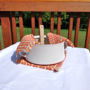 May include: White ceramic pot with a wooden spoon inside, sitting on a woven orange and beige pot holder. The pot holder is on a white tablecloth.