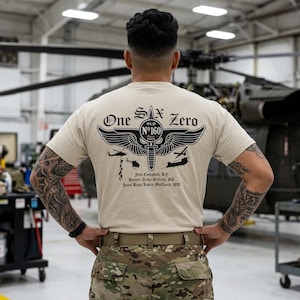May include: A beige t-shirt featuring a black graphic on the back. The design includes a central dagger with wings, the text "One Six Zero" and "Old No 160", and silhouettes of helicopters. Additional text lists "Fort Campbell, KY", "Hunter Army Airfield, GA", and "Joint Base Lewis McChord, WA".