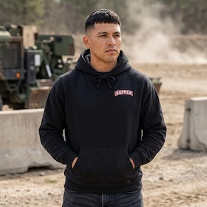 May include: A black hooded sweatshirt featuring a front kangaroo pocket and a rectangular patch on the chest with the word SAPPER printed in white letters on a red background. The hoodie includes matching black drawstrings at the neckline.