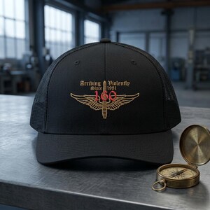 May include: A black trucker hat with a mesh back and a front panel featuring an embroidered gold winged sword emblem, the red number 160, and the text Arriving Violently Since 1981. A small, round, metallic brass-colored compass sits to the right.