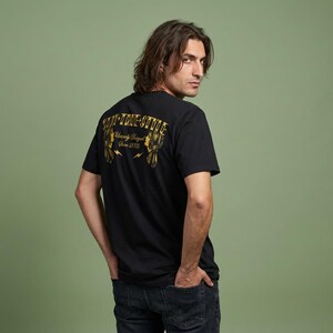 Op de afbeelding: Zwart T-shirt met gouden tekst op de rug: "Stay True Style" boven "Uniquely Inspired Since 2023". Het shirt heeft een ronde hals en korte mouwen. Het model draagt een donkerblauwe jeans.