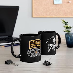 May include: Two black ceramic mugs. One mug features the words "RANGER AIRBORNE" in a yellow and white design with a parachute and wings. The other mug has a skeleton drinking from a cup with the words "LEG TEARS".