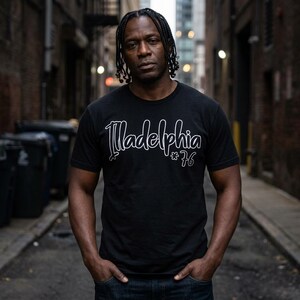May include: A black short-sleeved t-shirt featuring the white cursive text "Illadelphia" across the chest, followed by a small asterisk symbol and the number "76" in a white outline font. The shirt has a crew neckline and a standard fit.