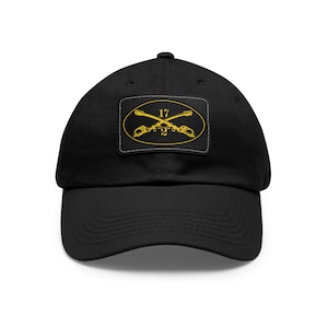May include: Black baseball cap with a curved brim. The cap features a rectangular patch with a gold emblem of crossed rifles and the number 17 inside an oval. The patch is outlined with a white border.