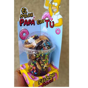 Puede incluir: Una taza de plástico transparente llena de mini donas, cubierta con glaseado de chocolate y chispas de colores. La taza está dentro de una caja amarilla con gráficos de donas rosas y el texto "Feliz Dia Para". Se ve una imagen de dibujos animados de Homer Simpson.